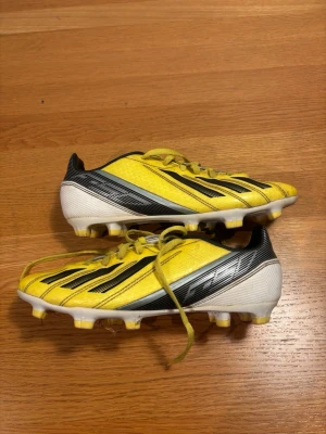 Gula Adidas f50 fotbollsskor  - Snygga gula fotbollsskor från Adidas med klassiska svarta ränder och vita detaljer. Skorna har snörning och fasta dobbar för bra grepp på planen. Tillverkade i syntetmaterial som ger lätt känsla och bra bollkontroll. Perfekta för dig som vill sticka ut på fotbollsplanen.