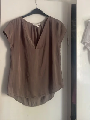 Beige v-ringad blus med kort ärm - Snygg brun/beige blus med djup v-ringning och korta ärmar. Blusen har en lätt glansig finish och rundad nederkant som är något längre bak. Den är i storlek 36 men passar även för större storlekar.