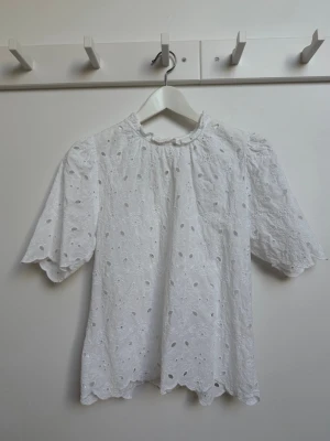 Vit blus med broderi Anglaise - Vit blus med broderi Anglaise från Lindex. Blusen har korta puffärmar och vågig kant nertill och i ärmsluten. Perfekt för en romantisk och somrig look. Tillverkad i ett lätt och luftigt bomullstyg, skön för varma dagar. Lapparna är borttagna då de syntes igenom.