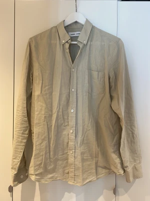 Beige linneskjorta från Samsøe Samsøe - Stilren beige skjorta från Samsøe Samsøe i svalt linne. Klassisk krage, bröstficka och vita knappar framtill. Perfekt för en avslappnad men ändå snygg look. Långärmad modell med normal passform.
