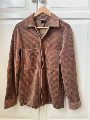 Brun manchester overshirt från H&M - Overshirt i brun manchester från H&M med två stora bröstfickor och tryckknappar framtill. Skjortan har klassisk krage och raka ärmar med knapp vid ärmslut. Perfekt för lager på lager och ger en avslappnad men snygg look.