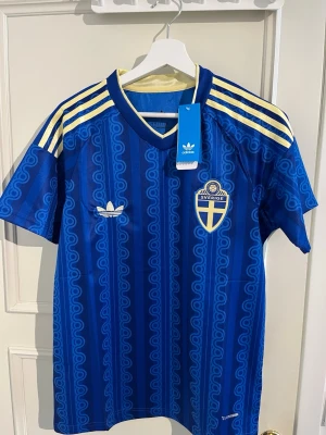 Blå Sverige tröja från Adidas - Säljer en blå Sverige matchtröja från Adidas med gula detaljer och klassiska ränder på axlarna. Tröjan har ett mönster i olika blå nyanser, broderad Sverige-logga och Adidas-märke på bröstet. Perfekt för dig som vill stötta landslaget med stil.