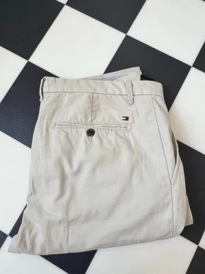 Beiga chinos från Tommy Hilfiger - Snygga beiga chinos från Tommy Hilfiger med klassisk passform. Byxorna har bälteshällor, knappstängning bakficka och den ikoniska Hilfiger-loggan broderad ovanför fickan. Tillverkade i mjukt bomullstyg som ger en clean och stilren look.