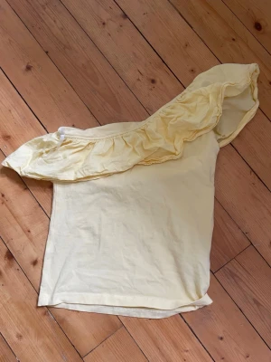 Ljusgul oneshoulder topp med volang - Snygg ljusgul oneshoulder topp med bred volang längs axeln. Toppen är ärmlös och har en feminin, somrig look. Perfekt för dig som vill sticka ut med en unik detalj. Tillverkad i mjukt material som känns skönt mot huden.