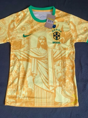 Gul Brasil fotbollströja Nike - Säljer en gul Brasilien fotbollströja från Nike med gröna detaljer och ett stort motiv av Kristusstatyn på framsidan. Tröjan har CBF-emblemet, fyra stjärnor och Nike-logga broderat på bröstet. Tillverkad i lätt och ventilerande material.