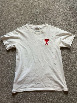 Vit t-shirt med rött hjärta från Ami - Vit t-shirt från Ami med ett litet rött hjärta och bokstaven A broderat på bröstet. Klassisk rund halsringning och korta ärmar. Tillverkad i mjuk bomull för en bekväm känsla. Perfekt till jeans eller shorts.