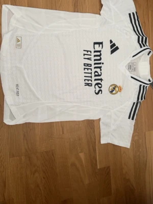 Real Madrid matchtröja Adidas vit - Säljer en vit Real Madrid matchtröja från Adidas med svarta detaljer och klubbmärke på bröstet. Tröjan har korta ärmar, rund hals och tre svarta ränder på axlarna. Tillverkad i lätt och ventilerande material med HEAT.RDY-teknologi.