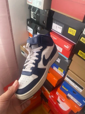 Nike Court Borough Mid 2 sneakers - Snygga sneakers från Nike i blått och vitt med klassisk swoosh-logga på sidan och Nike-tryck på kardborrebandet. Skorna har en mid cut-design, vita skosnören och perforerad tå för extra ventilation. Perfekta för dig som gillar sportig och stilren look.