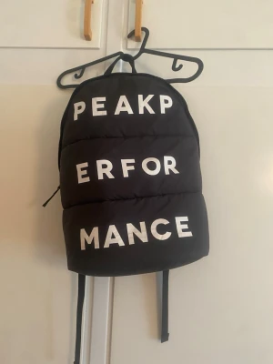Svart Peak Performance ryggsäck - Snygg svart ryggsäck från Peak Performance med stora vita bokstäver på framsidan. Ryggsäcken har vadderad design, justerbara axelremmar och dragkedja upptill. Perfekt för skolan eller träningen.
