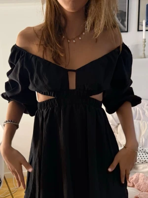 Somrig cutout klänning  - Trendig svart kortklänning med offshoulder-design och trekvartsärmar. Klänningen har snygga cut-outs i midjan och en v-ringad urringning med liten detalj framtill. Perfekt för dig som vill ha en modern och stilren look.