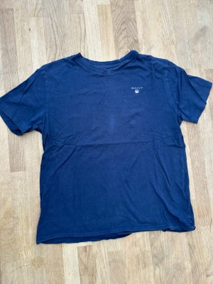 Mörkblå t-shirt från GANT - En klassisk mörkblå t-shirt från GANT med liten logga och sköld på bröstet. T-shirten har rund halsringning och korta ärmar. Tillverkad i mjuk bomull för en bekväm känsla och enkel stil.
