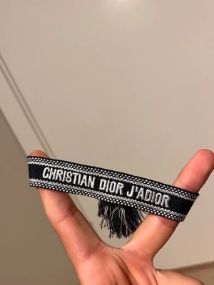 Christian Dior J'Adior armband svart - Snyggt svart armband från Christian Dior med texten 'J'Adior' broderad i vitt. Armbandet är gjort i tyg och har dekorativa tofsar i ändarna. Perfekt accessoar för att lyfta din outfit med en exklusiv touch.