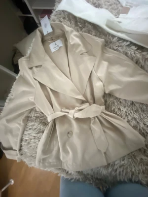Beige trenchcoat med bälte - Stilren beige trenchcoat med bred krage och knytskärp i midjan. Jackan har knappar framtill och en klassisk, avslappnad passform. Ifrån stradivarius och i storlek S. Helt ny med lapp på men finns en liten defekt som syns inte men se i bild 5💗💗