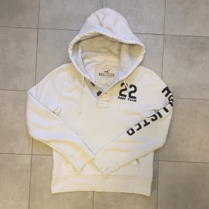 Vit Hollister  - Vit sällsynt hoodie från Hollister med svart tryck på ärmen och bröstet där det står '22 Surf Team'. Hoodien har en stor huva med dragsko och ribbade muddar vid ärmar och nederkant. Perfekt för en avslappnad och sportig stil. Har inga hål eller fläckar. Skriv gärna vid frågor och annat🙂