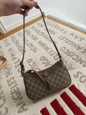 Gucci axelväska i beige monogram - Snygg axelväska från Gucci i beige canvas med klassiskt GG-monogram och bruna läderdetaljer. Väskan har guldfärgad logga framtill, justerbar axelrem och dragkedja upptill. Perfekt storlek för det viktigaste och ger en lyxig touch till din outfit.