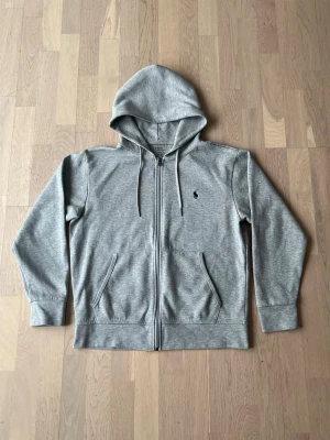 Grå zip hoodie från Polo Ralph Lauren - Snygg grå zip hoodie från Polo Ralph Lauren med klassisk svart logga broderad på bröstet. Hoodien har dragsko i huvan, två fickor framtill och ribbade muddar vid ärmar och nederkant. Tillverkad i mjuk bomullsblandning för skön komfort.