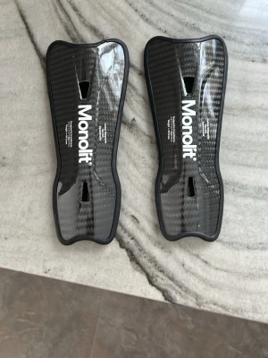 Monolit benskydd i kolfiber - Säljer ett par svarta benskydd från Monolit i kolfiber med ergonomisk form och vadderad insida för extra komfort. Lätta och slitstarka, perfekta för fotboll eller andra sporter där skydd behövs. Snygg design med Monolit-logga på framsidan. Väger endast 40 gram för extra smidighet.