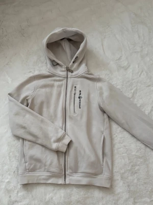 Vit hoodie från Sail Racing - Vit hoodie från Sail Racing med dragkedja framtill och en bröstficka med logga. Hoodien har justerbar huva med snören och ribbade muddar vid ärmslut och nederkant. Storlek 150, passar som xxs 