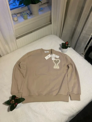 Beige sweatshirt från Ami Paris - Säljer en beige sweatshirt från Ami Paris med deras ikoniska hjärta-A-logga broderad på bröstet. Tröjan har rund hals, ribbade muddar och är i mjukt bomullsmaterial. Perfekt för en clean och stilren look.