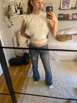 Vit spetsblus med volangdetaljer - Säljer en superfin vit blus i spets med volangdetaljer på axlarna och runt ärmhålen. Toppen är croppad och har en hög hals med fina broderade mönster. Perfekt att styla med jeans eller kjol för en trendig look.