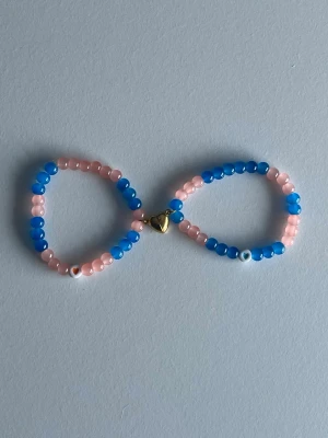 Blått och rosa pärlarmband med magnet-hjärta - Sött armband med runda pärlor i blått och ljusrosa, dekorerat med ett guldfärgat hjärtlås. Armbandet har även små vita och orange detaljer. Perfekt accessoar för att addera färg till din stil.