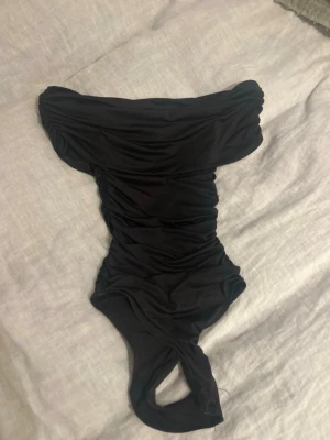 Svart offshoulder body - Trendig svart offshoulder body med rynkade detaljer och croppad passform. Perfekt för dig som vill ha en stilren och edgy look. Toppens material känns mjukt och följsamt mot kroppen. Litet hål på sidan men syns knappt