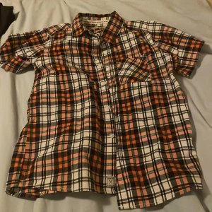 Rutig kortärmad skjorta från Denim & Co - Säljer en rutig skjorta från Denim & Co med korta ärmar. Skjortan har en klassisk krage, bröstficka och knappar framtill. Färgerna är orange, svart, vit och lite grönt. Perfekt för dig som gillar en avslappnad men ändå snygg stil.