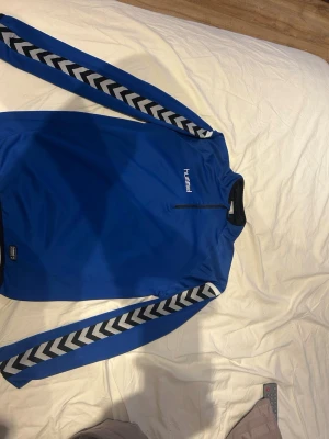 Blå träningsjacka från Hummel - Snygg blå träningsjacka från Hummel med dragkedja vid halsen och klassiska vita och svarta chevronränder längs ärmarna. Tillverkad i ett mjukt och stretchigt funktionsmaterial som passar perfekt till träning eller uppvärmning.