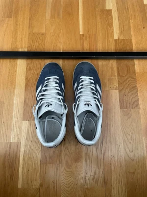 Adidas Originals gazzel blåvita sneakers - Mörkblå sneakers från Adidas gazzel  Originals med klassiska vita ränder på sidorna och vit sula. Ovandelen är i mocka och skorna har vita skosnören samt detaljer i vitt på häl och tunga. Perfekta för en sportig och stilren look. Lite slitna men inget man tänker på så! 
