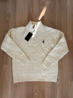 Vit kabelstickad tröja - Kabelstickad vit tröja med halv dragkedja och ribbad hög krage. Klassisk marinblå logga broderad på bröstet. Tröjan har långa ärmar och ribbade muddar. Perfekt för lager på lager och en clean look.