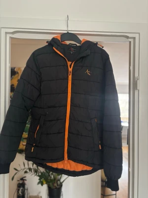 Svart pufferjacka med orange detaljer - Snygg svart pufferjacka med orange dragkedja och foder. Jackan har huva, två sidofickor med dragkedja och quiltad design. Perfekt för kalla dagar och riktigt skön att bära. Märke: Active by Tchibo. Materialet är syntet och jackan är vadderad. Slitet på loggan. 