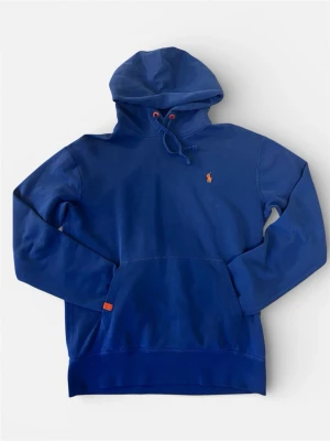 Blå hoodie från Polo Ralph Lauren - Snygg blå hoodie från Polo Ralph Lauren med klassisk huva och dragsko. Ikoniska lilla polospelaren broderad i orange på bröstet och en stor magficka framtill. Tillverkad i mjuk bomullsblandning och har ribbade muddar vid ärmslut och nederkant.