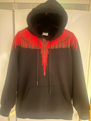 Svart hoodie med röd fjäderdetalj - Säljer en svart hoodie från Marcelo Burlon med ikoniskt rött fjädermönster över axlar och bröst. Hoodien har stor huva, dragsnören och en klassisk magficka. Perfekt för dig som vill sticka ut med en unik och trendig design.