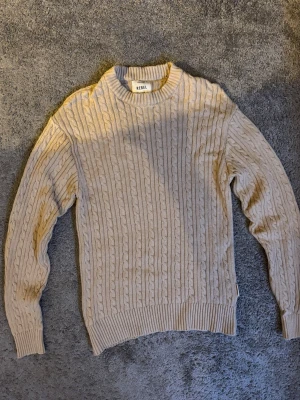 Beige kabelstickad tröja Rebel XS - Snygg beige kabelstickad tröja från Rebel i storlek XS. Tröjan har rund halsringning, ribbade muddar och nederkant samt ett klassiskt kabelmönster. Perfekt för lager på lager och enkel att matcha med olika outfits.