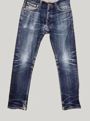 Mörkblå jeans från Nudie Jeans - Snygga mörkblå jeans från Nudie Jeans med slitna detaljer och kontrastsömmar. Klassisk femficksmodell med raka ben och coola whiskers-effekter på både fram- och baksida. Tillverkade i bomull för en bekväm känsla och tidlös look.