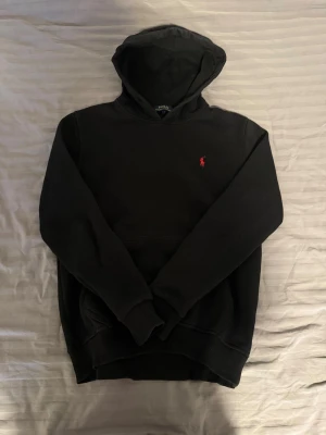 Svart hoodie från Ralph Lauren - Klassisk svart hoodie från Ralph Lauren med röd broderad logga på bröstet. Hoodien har huva, magficka och ribbade muddar vid ärmar och nederkant. Tillverkad i mjukt bomullsmaterial som är skönt att bära.