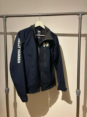 Marinblå Helly Hansen vindjacka - Snygg marinblå vindjacka från Helly Hansen med dragkedja och hög krage. Jackan har vit logga på bröstet och ärmen samt en bröstficka med dragkedja. Perfekt för blåsiga dagar och enkel att matcha med olika outfits.                                                                   Skriv DM vid intresse🙌🏽