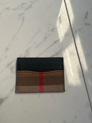 Burberry kortfodral i läder och canvas - Snyggt kortfodral från Burberry i svart läder och klassisk beige- och brunrutig canvas med en röd rand i mitten. Perfekt för kort och ID, smidig att ha i fickan. Stilren design med flera kortfack och exklusiv känsla.