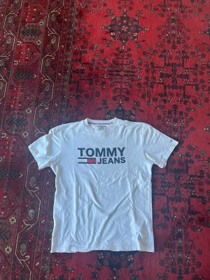 Vit T-shirt från Tommy Hilfiger - Säljer en klassisk vit T-shirt från Tommy Jeans med stor logga framtill i blått och rött. T-shirten har rund halsringning och korta ärmar. Tillverkad i mjuk bomull för skön känsla och enkel stil.