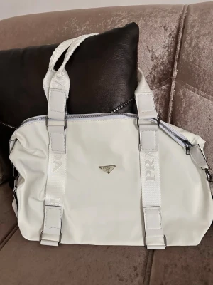 Vit handväska från Prada - Snygg vit handväska från Prada med silverfärgad dragkedja och detaljer. Väskan har breda axelband med logotyp och en stilren metallogo framtill. Perfekt för dig som gillar en clean och modern look. Dom är inte använda alls och stick 250 