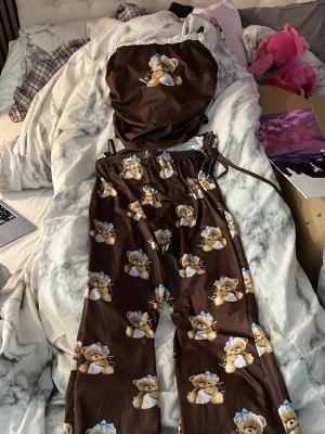 Brun pyjamas med nallemönster - Söt brun pyjamas med matchande linne och byxor, täckt av gulliga nallar med blå mössa. Perfekt för mysiga kvällar hemma. Byxorna har resår i midjan och linnet har tunna axelband. Använd 1 gång. Vid intresse skicka dm.