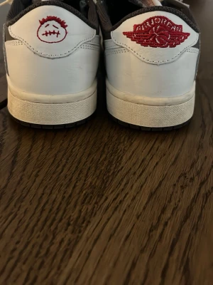 Nike Air Jordan 1 Low Travis Scott - Nike Air Jordan 1 Low Travis Scott i vitt och mörkbrunt läder med ikoniska röda detaljer på hälen. Snygg kontrast mellan den vita ovandelen och den mörka tån. Klassisk Nike Air-logga på plösen och Travis Scott smiley på baksidan.