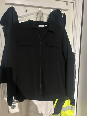 Svart overshirt med dragkedja - Stilren svart overshirt med dragkedja framtill och två bröstfickor med lock. Klassisk krage och raka ärmar. Perfekt att bära som lager över t-shirt eller hoodie. Materialet ser ut att vara mjukt och något tjockare, vilket ger en schysst look.