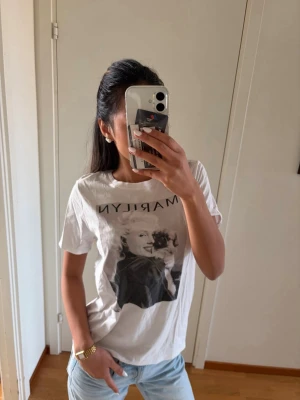 Vit Marilyn Monroe t-shirt - Säljer en vit t-shirt med tryck av Marilyn Monroe på framsidan. T-shirten har rund hals och korta ärmar, samt en avslappnad passform. Perfekt för dig som gillar ikoniska motiv och vill ha en cool, casual stil.