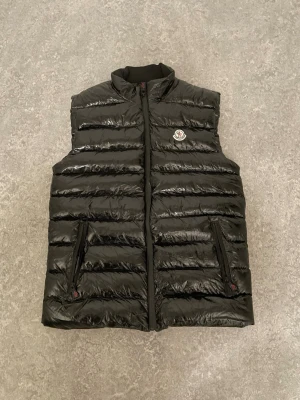Svart Moncler väst - Snygg svart dunväst från Moncler med glansig finish och quiltad design. Västen har två dragkedjeförsedda fickor fram, hög krage och ett Moncler-märke på bröstet. Insidan har en röd detaljficka och ett serietidningsmönster. Perfekt för lager-på-lager-stil.
