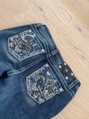 Mörkblå Miss Me jeans - Säljer ett par mörkblå jeans från Miss Me med snygga broderade bakfickor och dekorativa nitar. Jeansen har kontrastsömmar, bootcut-modell och slitningar framtill för en cool look. Perfekta för dig som vill sticka ut med detaljer!