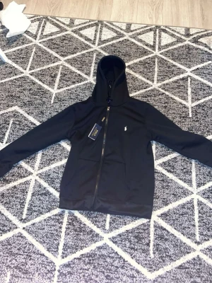 Svart hoodie från Polo Ralph Lauren - Snygg svart hoodie från Polo Ralph Lauren med dragkedja framtill och klassisk huva. Ikoniska lilla loggan broderad på bröstet. Tillverkad i mjukt material som passar perfekt till en avslappnad stil. Långa ärmar och fickor framtill.