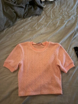 Rosa stickad topp från Zara - Säljer en söt ljusrosa stickad topp från Zara med korta ärmar och rund halsringning. Tröjan har ribbade muddar vid hals och ärmslut samt ett diskret kabelstickat mönster. Perfekt för dig som gillar en mjuk och trendig look. Storlek M men är liten i storleken 