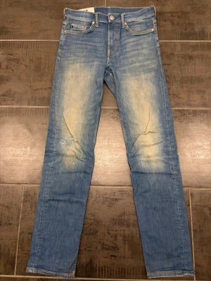 Slim fit blå jeans från &Denim - Snygga slim fit jeans i blå denim från &Denim. Jeansen har slitningar och ljusa partier på låren för en trendig, använd look. Klassisk femficksmodell med knappgylf och smal passform. Perfekta till vardagsstilen. Storlek 30/34