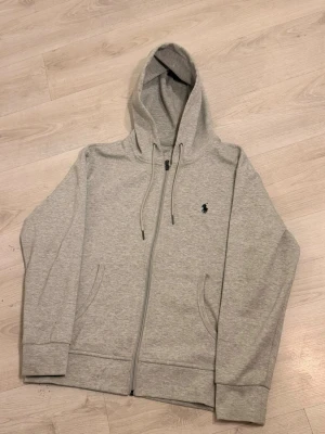 Grå zip hoodie från Polo Ralph Lauren - Snygg gråmelerad hoodie med dragkedja från Polo Ralph Lauren. Klassisk huva med dragsko, två fickor framtill och ikoniska broderade loggan på bröstet. Perfekt för en chill och stilren look. Mjuk bomullsblandning som känns skön mot huden.